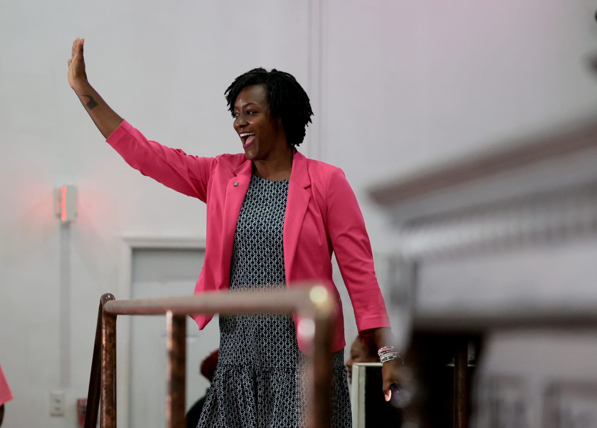 A smiling woman in a bright pink blazer and patterned dress waves her raised hand while standing indoors near a railing.