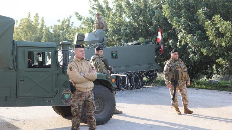 Soldiers in camouflage uniforms stand near a green military Humvee and an armored vehicle, with barbed wire and a Lebanese flag visible in the background.