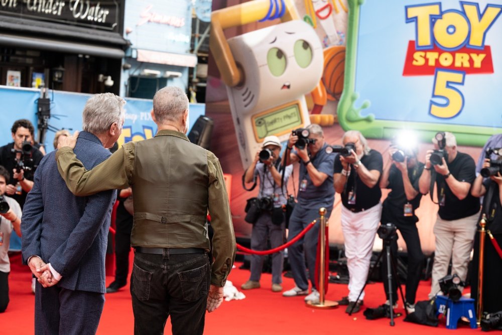 Two men stand on a red carpet posing for a crowd of photographers, with a large "Toy Story 5" backdrop featuring a Forky character balloon displayed behind them.