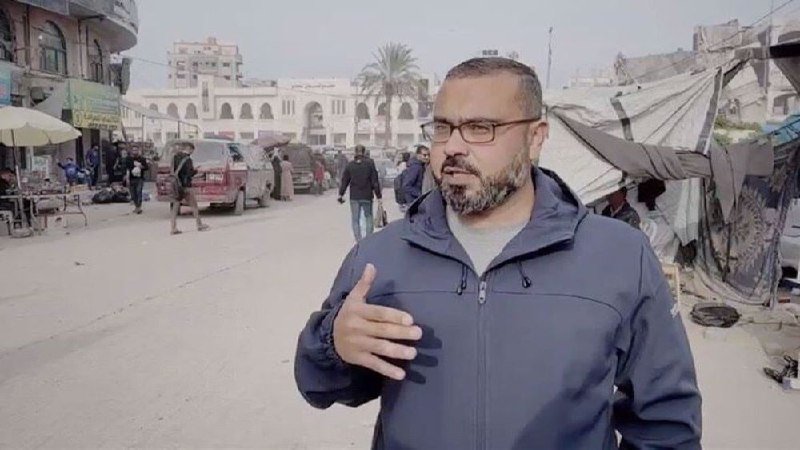 A bearded man in a blue jacket gestures with his hand while speaking on a street with pedestrians, vehicles, damaged buildings, and tents in the background.