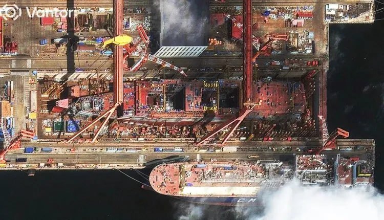A satellite image shows a large red and white ship docked at an industrial port, surrounded by cranes, construction equipment, and structures emitting white smoke or steam.