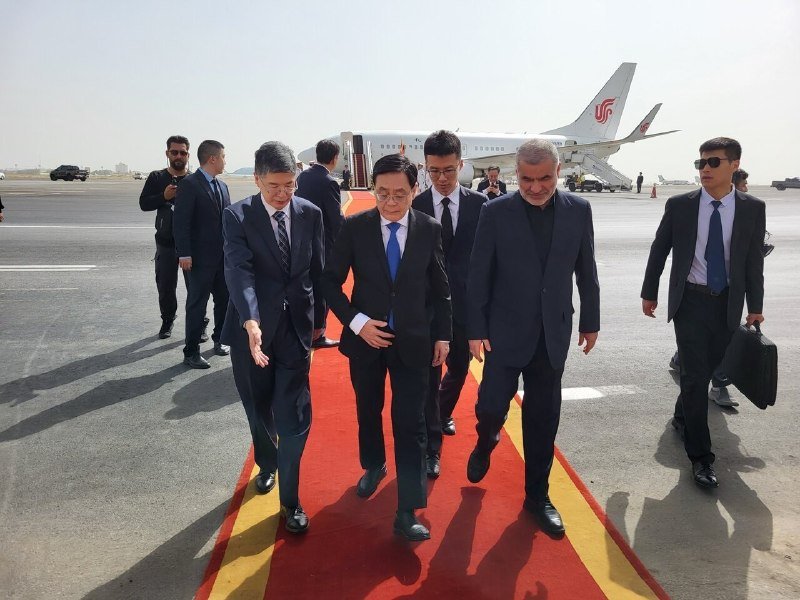 A group of men in dark suits walk along a red carpet on an airport tarmac, with a commercial airplane and mobile staircase visible in the background.
