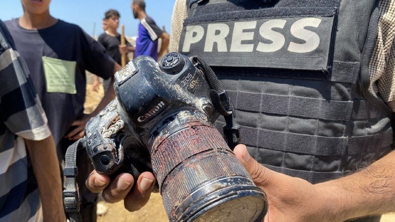 A person wearing a vest labeled "PRESS" holds a dust-covered Canon DSLR camera, while other people stand in the sunny, sandy background.