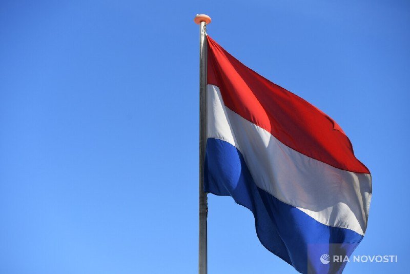 A red, white, and blue horizontal-striped flag waves on a flagpole against a clear blue sky, bearing a "RIA Novosti" watermark.