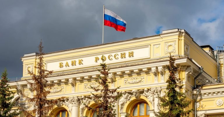 The Russian flag flies atop a yellow neoclassical building inscribed with "БАНК РОССИИ" against a cloudy sky, flanked by evergreen trees.