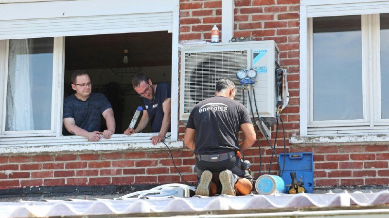 A technician wearing an "energies" branded shirt kneels outside, servicing a Daikin air conditioning unit while two men watch from an open window.