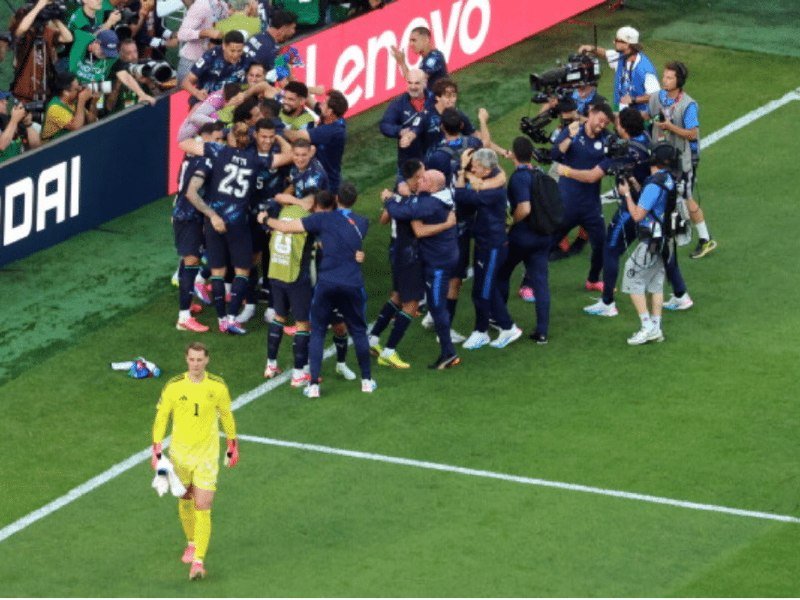 Soccer players in dark blue jerseys celebrate together in a huddle on the field while a goalkeeper in yellow walks away and cameramen film the scene.