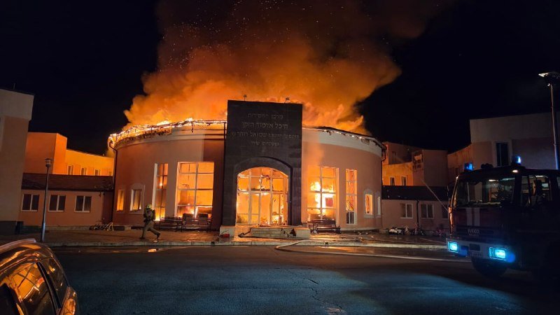 A building with a domed roof and arched entrance is engulfed in flames at night, with thick smoke rising into the sky as a fire truck and a person stand nearby.