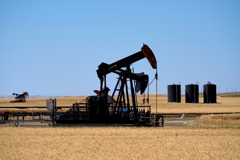 A pumpjack operates in a golden field under a clear blue sky, with oil storage tanks visible in the background.