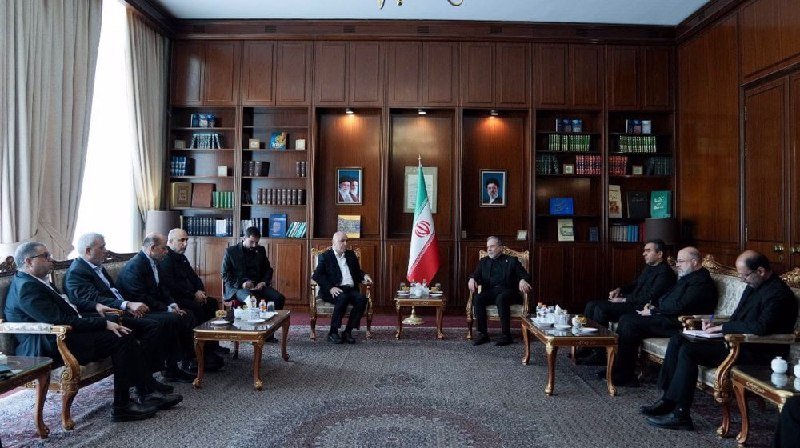 Men in suits sit in a wood-paneled room facing each other across small tables, with an Iranian flag and framed portraits displayed on the back wall.