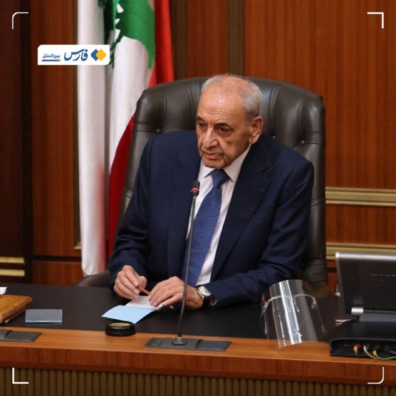 An elderly man in a dark suit sits at a wooden desk with a microphone, papers, and a face shield, before a Lebanese flag.