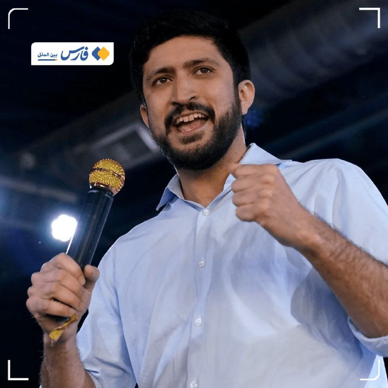 A bearded man in a light blue shirt speaks into a microphone, gesturing with his fist, with a logo reading "بين الملل" in the upper left corner.
