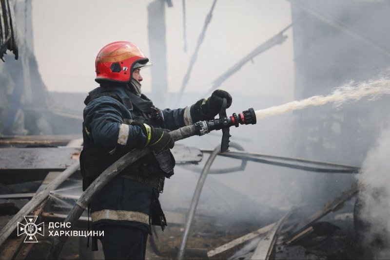 A firefighter in a red helmet and protective gear sprays water from a hose amid smoke and debris at a fire site, with a "ДСНС ХАРКІВЩИНИ" logo visible.