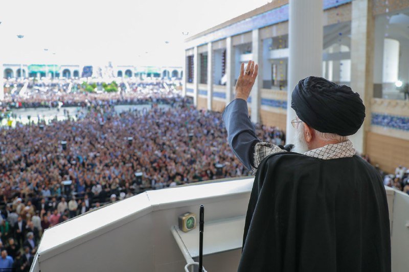 A bearded cleric in a black turban and robes stands at an elevated podium, raising his hand while addressing a massive crowd gathered in a large courtyard.