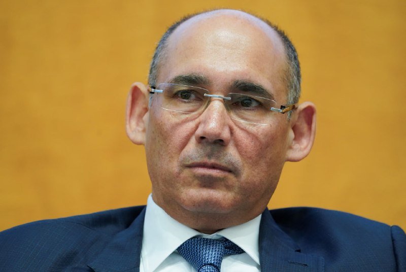 A man in a blue suit and tie with glasses looks forward against a wooden background.