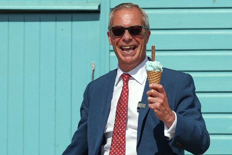 A smiling man in sunglasses, a blue suit, and a red patterned tie holds up a mint-green ice cream cone in front of a turquoise wooden wall.