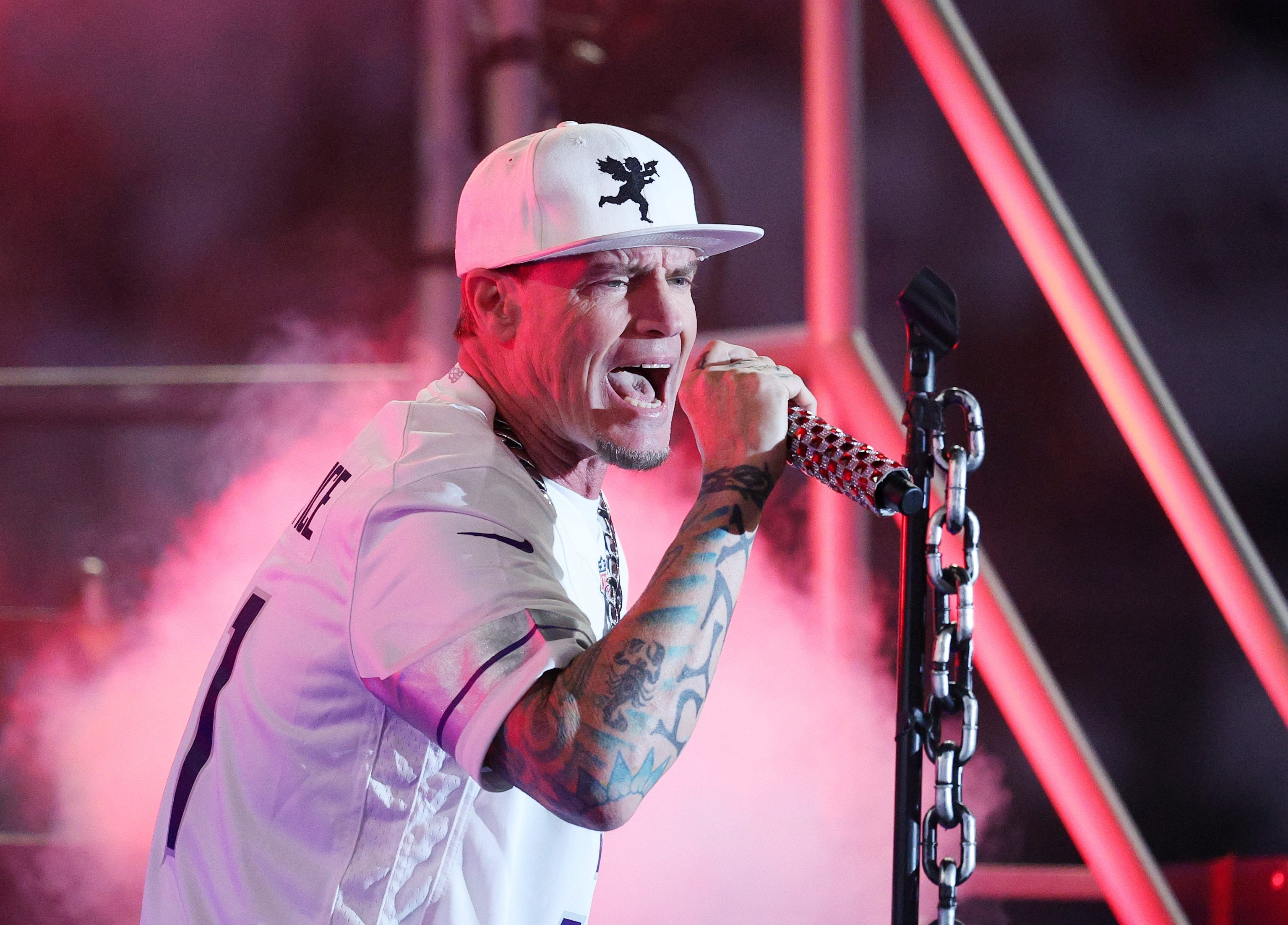 A tattooed man wearing a white cap and jersey sings passionately into a microphone on a red-lit stage, with chains visible on the mic stand.