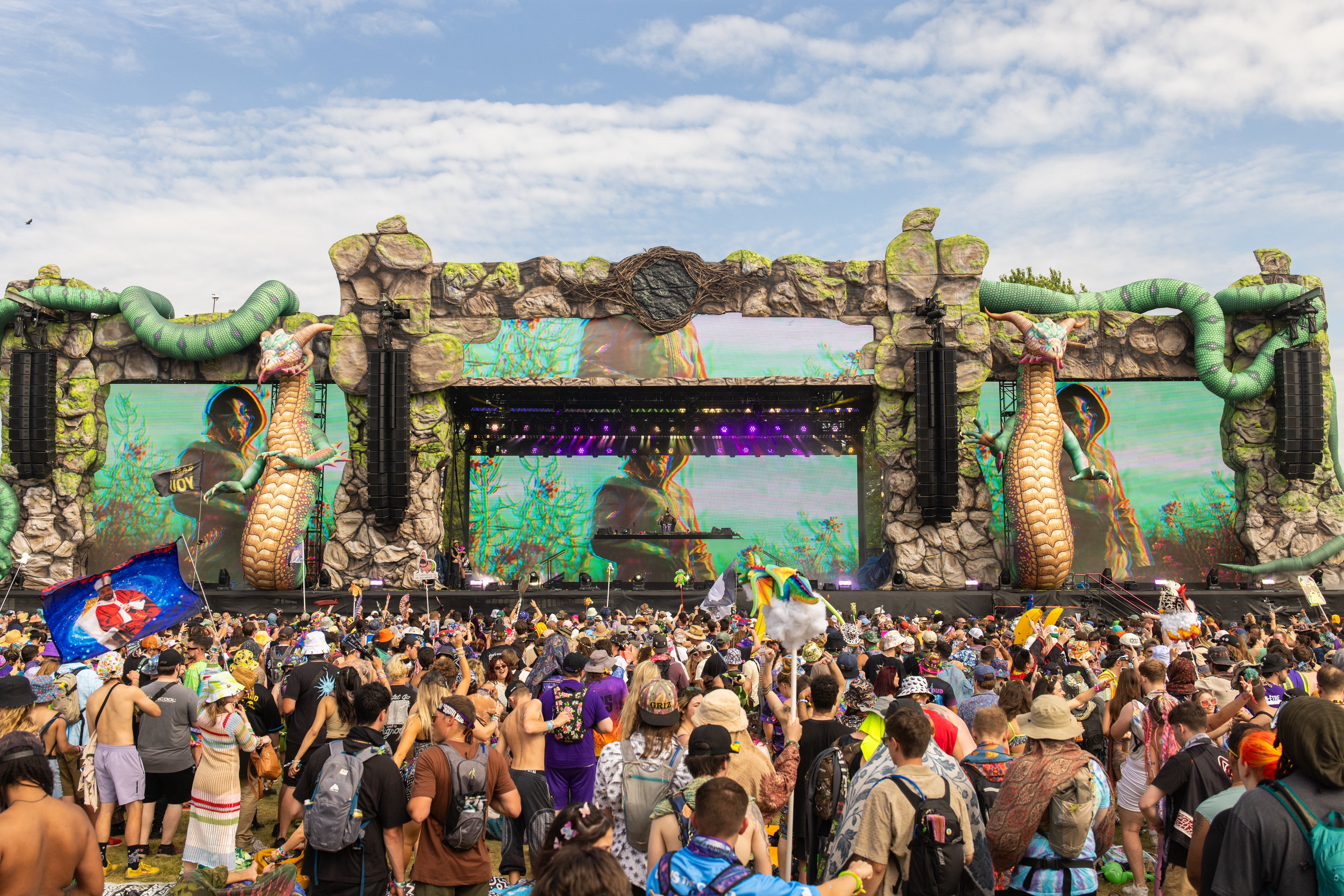 A large crowd gathers before an elaborate outdoor music festival stage featuring massive green serpent sculptures, rocky facades, and LED screens displaying colorful visuals under a partly cloudy sky.