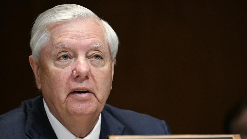 An older man with white hair, wearing a dark suit and white shirt, appears to be speaking against a dark background.
