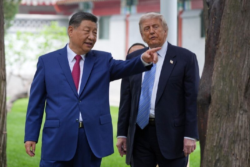 Two men in dark suits walk together outdoors on a lawn, one pointing forward, with a traditional pavilion visible in the background.