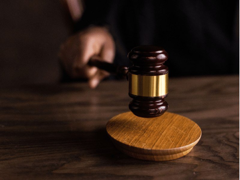 A hand grips a wooden gavel with a gold band, striking it against a circular wooden sound block on a dark wooden surface.