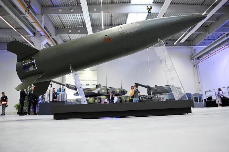 A large green bomb is displayed on angled supports inside a hangar-like exhibition hall, with military vehicles and visitors visible in the background.