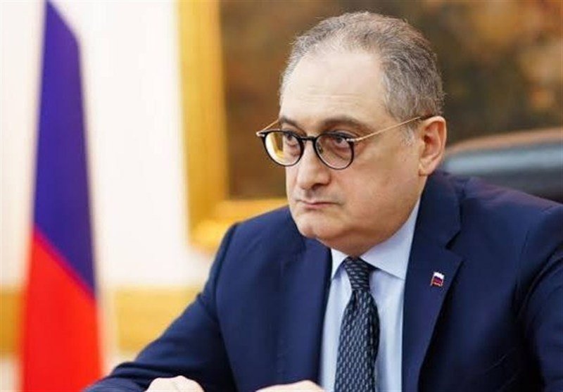 A bespectacled man in a dark suit and tie sits at a desk with a Russian flag visible behind him.