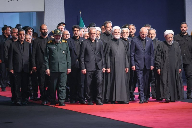 A group of men in dark suits, military uniforms, and clerical robes walk together along a red carpet.
