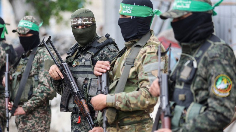 Masked militants in camouflage uniforms, green headbands, and face coverings stand together holding assault rifles.