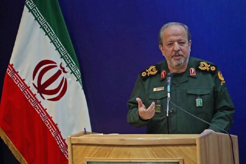 A bearded man in a green military uniform with insignia speaks at a wooden podium, gesturing with his hand, beside an Iranian flag.