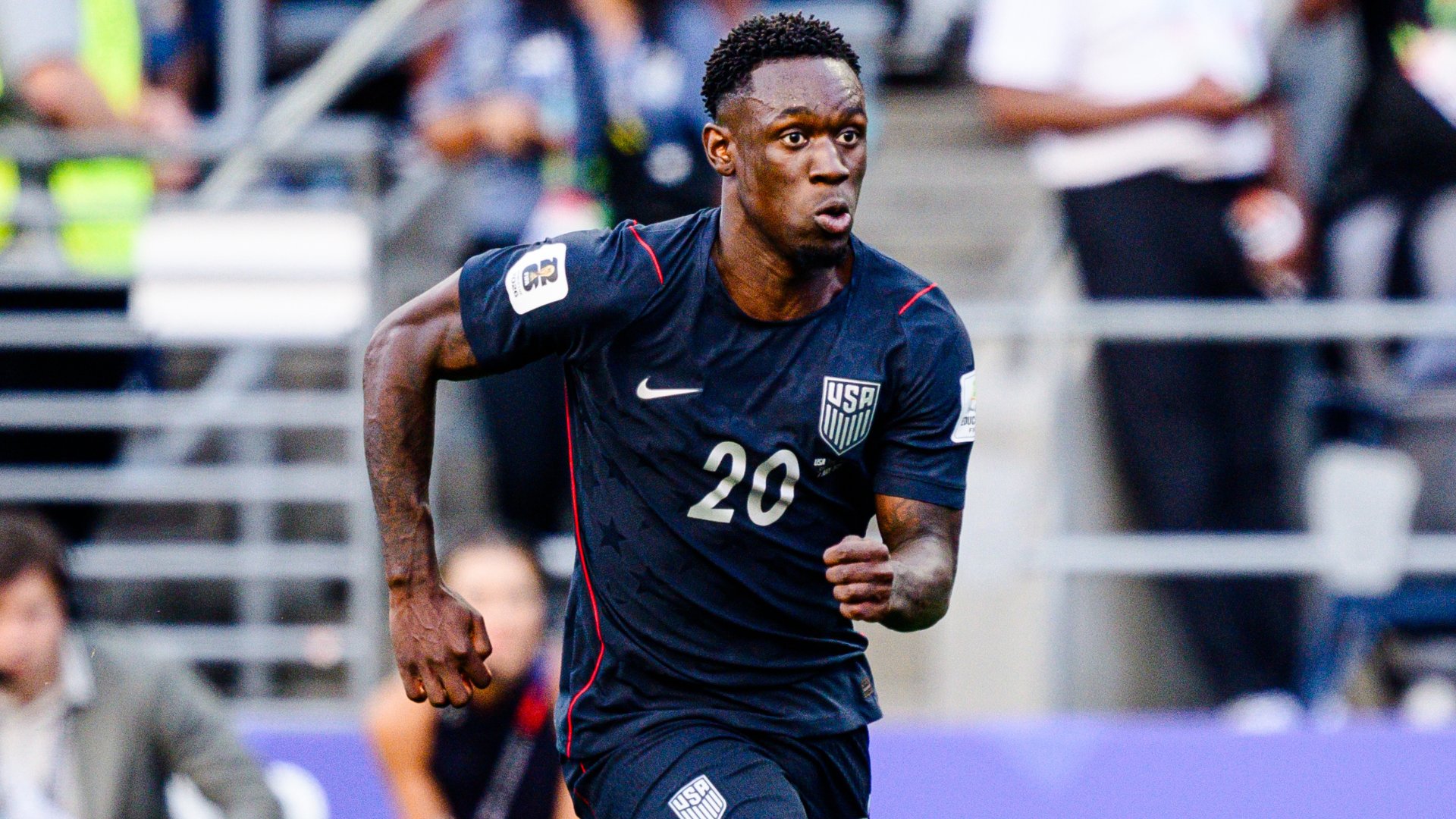 A soccer player wearing a dark blue USA jersey with the number 20 runs across a stadium field with blurred spectators in the background.