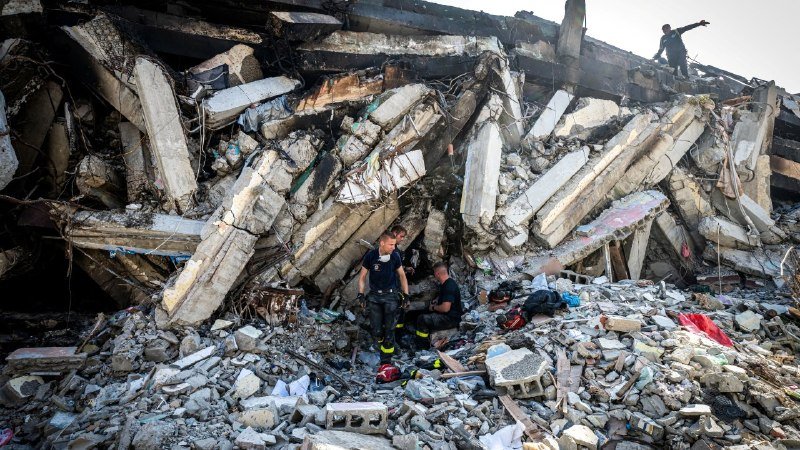 Rescue workers in dark uniforms search through the rubble of a collapsed concrete building.