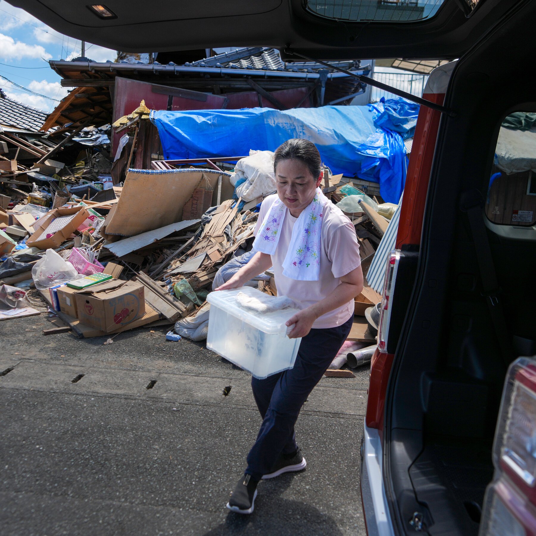 A woman carries a clear plastic storage bin away from a large pile of debris and damaged building materials toward an open vehicle trunk.