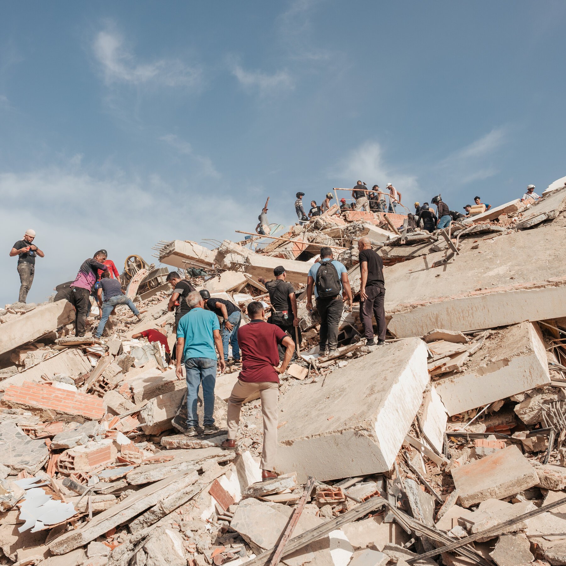 A coastal city shows widespread destruction, with collapsed multi-story buildings, debris-filled streets, vehicles, and distant intact high-rises along a shoreline.