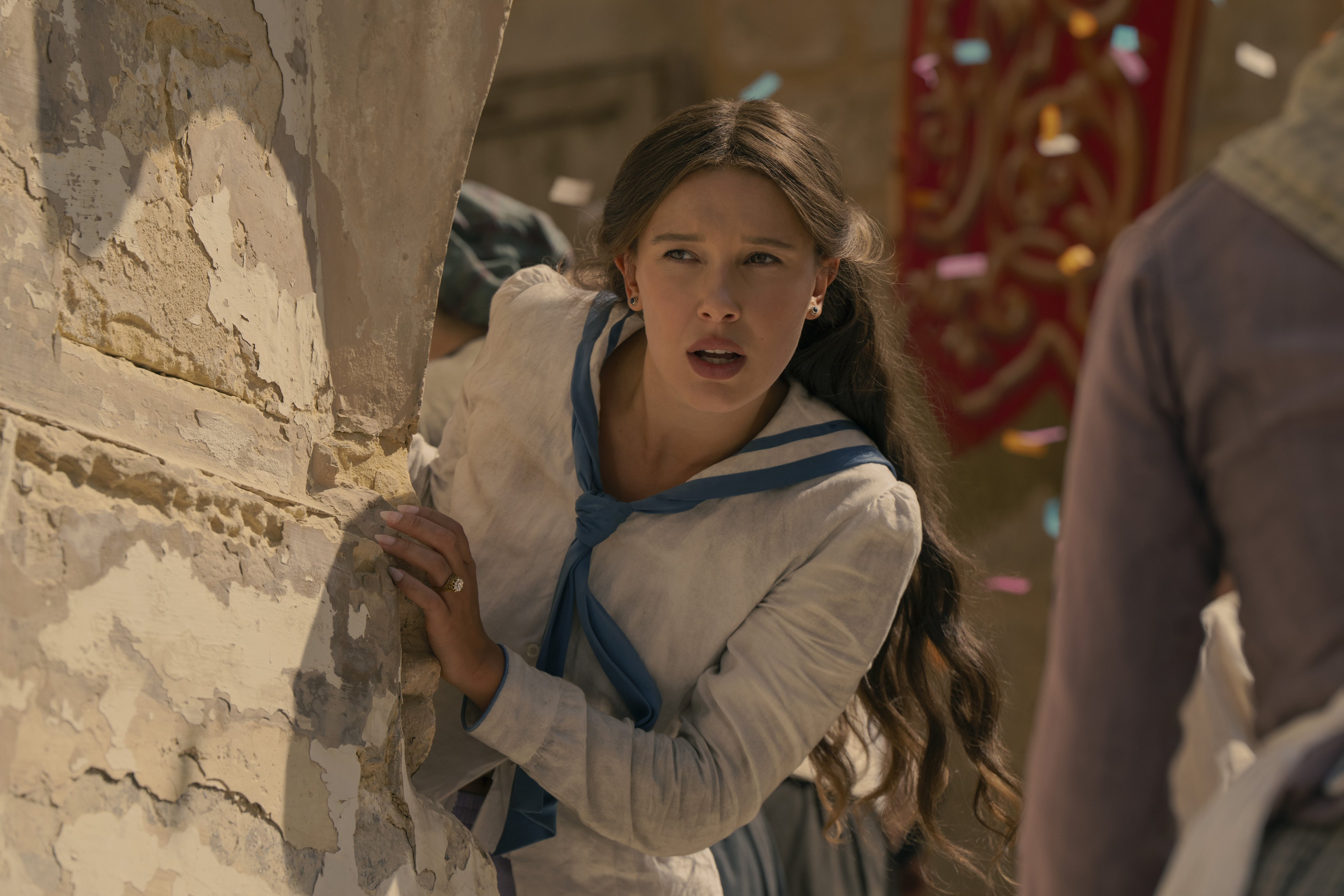 A woman with long brown hair, wearing a beige dress with a blue scarf, peeks cautiously from behind a weathered, peeling stone wall, with confetti falling in the background.