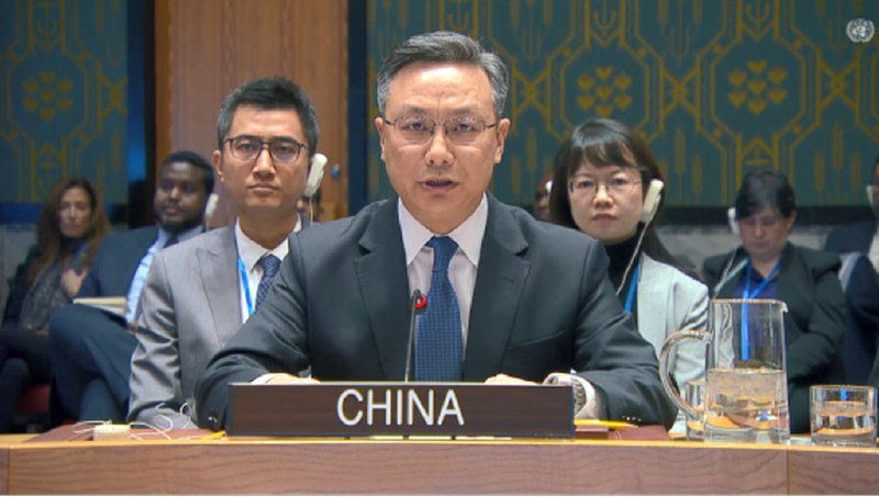 A man in a suit and glasses speaks at a desk bearing a "CHINA" placard, with other attendees seated behind him wearing headsets.