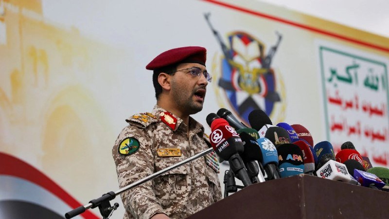 A military officer in a desert camouflage uniform and red beret speaks at a podium lined with numerous press microphones, with an emblem featuring crossed swords visible on the backdrop behind him.