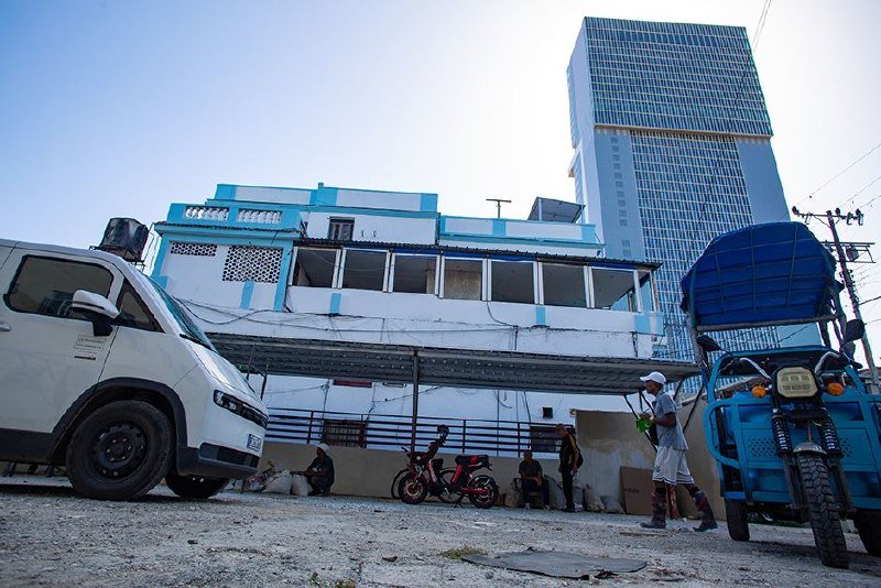 A white van, motorcycles, and a blue auto-rickshaw are parked near a blue and white building, with several people and a tall modern tower visible in the background.