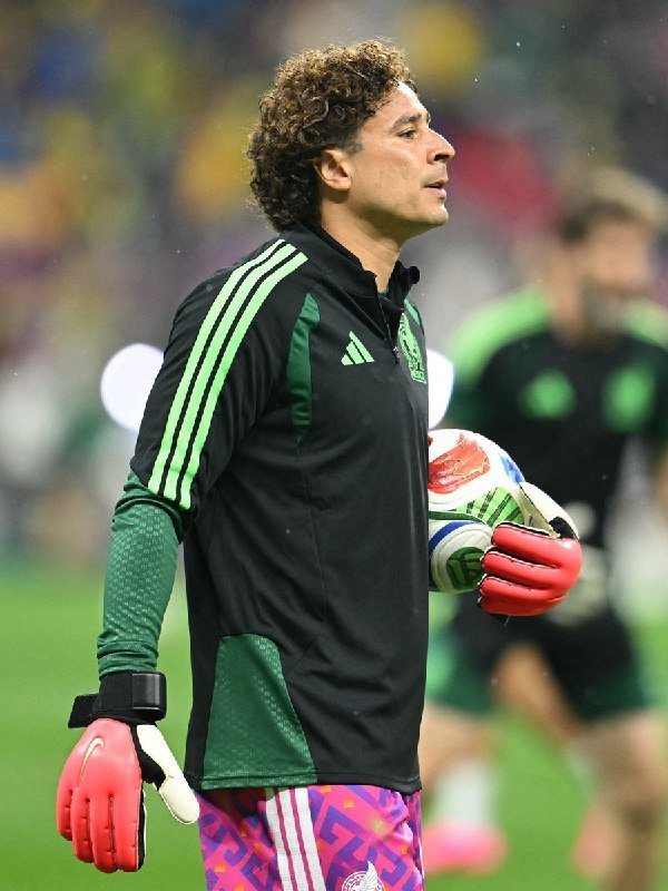 A curly-haired goalkeeper in a black and green training jacket, pink and purple patterned shorts, and red gloves holds a soccer ball during a pre-match warm-up.