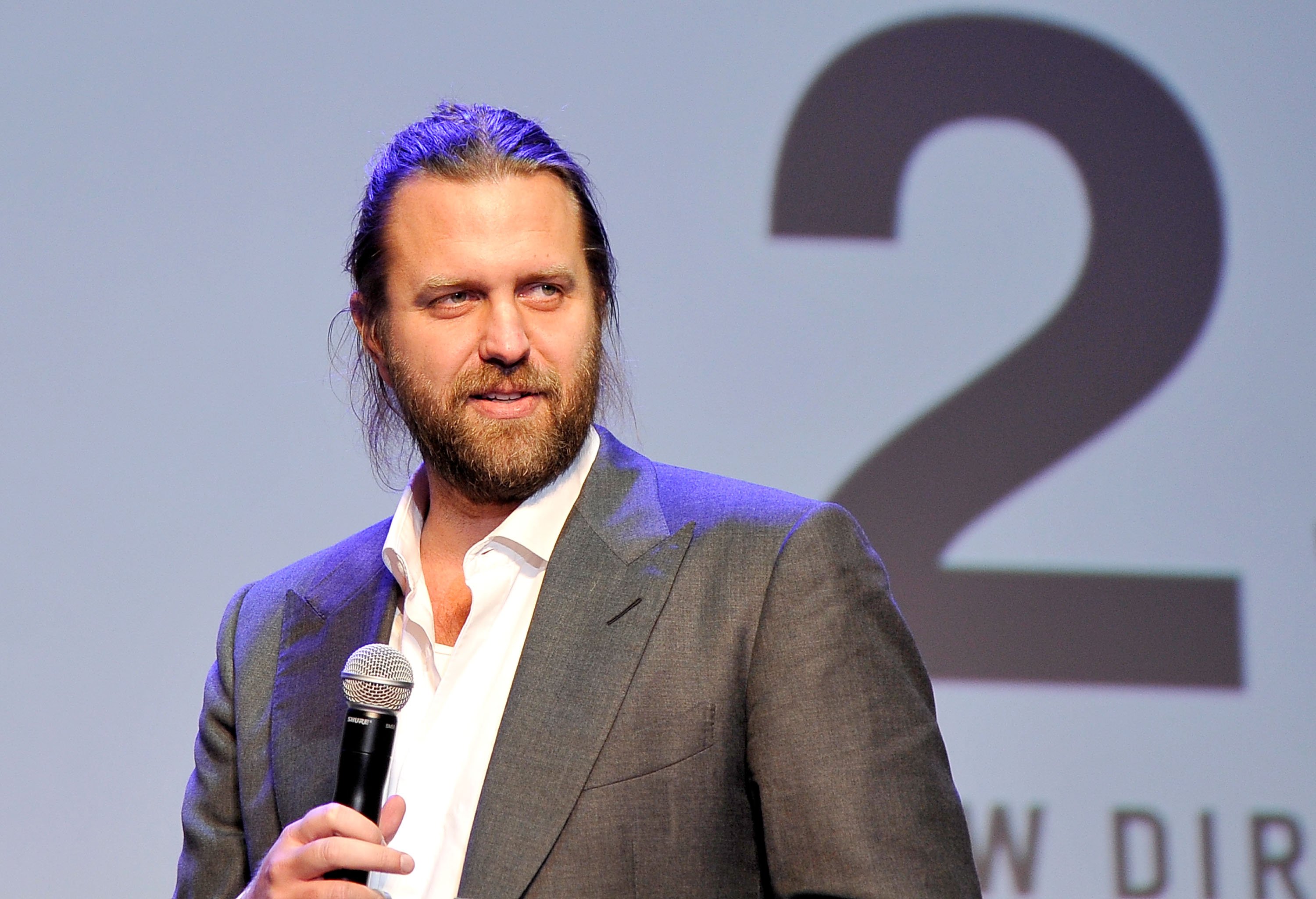 A bearded man with long hair, wearing a gray suit jacket and white shirt, holds a microphone while standing in front of a backdrop displaying a large number "2."