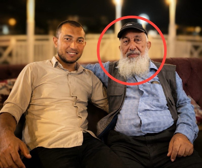 Two men sit together on a couch, with a younger man smiling on the left and an older man with a long white beard and dark cap on the right, whose face is circled in red.
