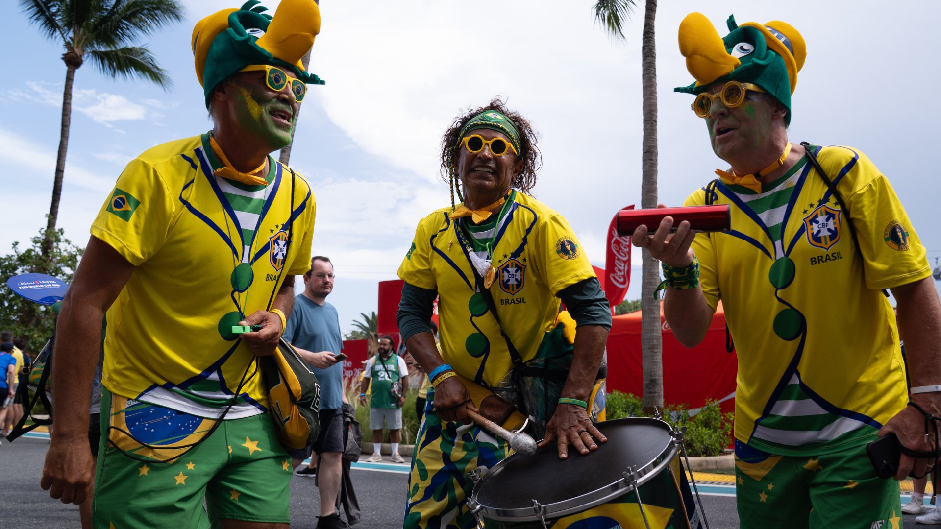 Three men in yellow Brazil jerseys and large green-and-yellow hats stand outdoors near palm trees, with one playing a drum and another holding a red noisemaker.