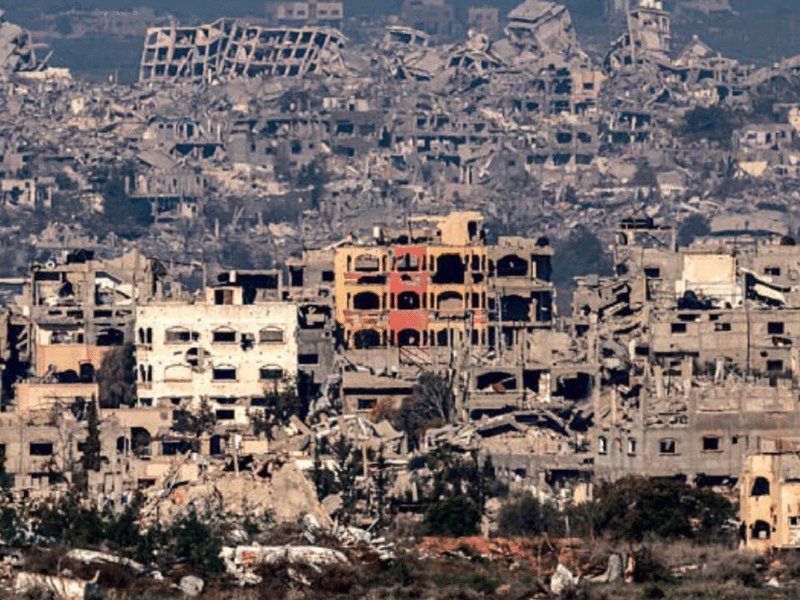 Extensive building rubble and heavily damaged multi-story structures stretch across a hillside, with only a partially intact red and beige building visible among the destruction.