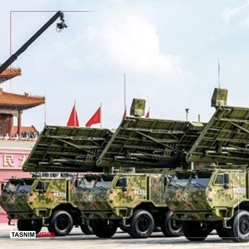 A row of camouflaged military missile launcher vehicles drive in formation during a parade, with red flags and a traditional building visible in the background.