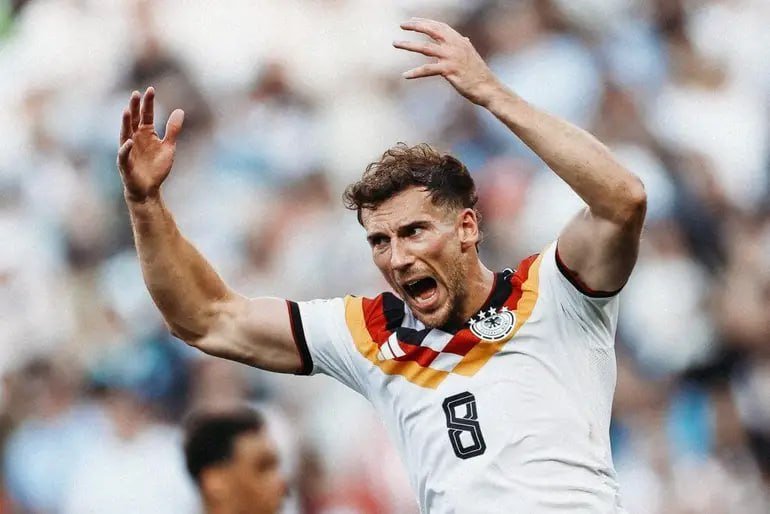 A soccer player wearing a white Germany jersey with the number 8 raises both arms and shouts, appearing frustrated, in front of a blurred stadium crowd.