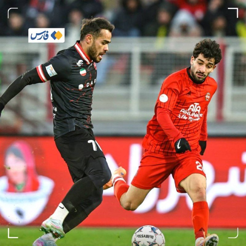 Two soccer players, one in a black uniform and one in red, compete for the ball during a match on a grass field.