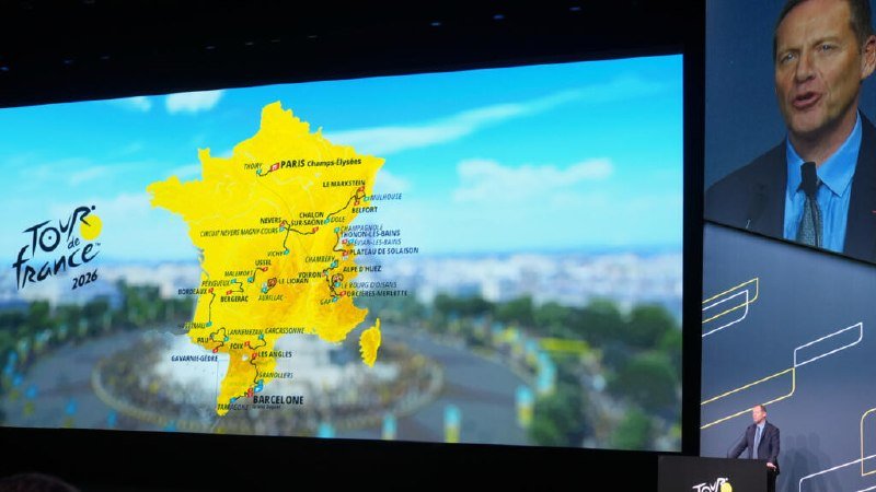 A man in a suit speaks at a podium beside a large screen displaying a yellow map of France marked with Tour de France 2026 cities and stage locations.