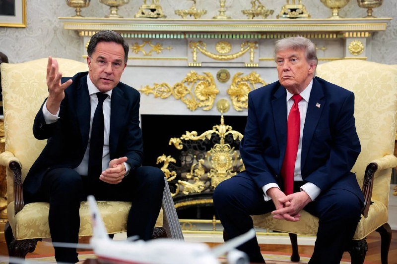 Two men in dark suits sit in ornate yellow chairs in a gilded room, one gesturing with his hand while speaking, the other listening with hands clasped.