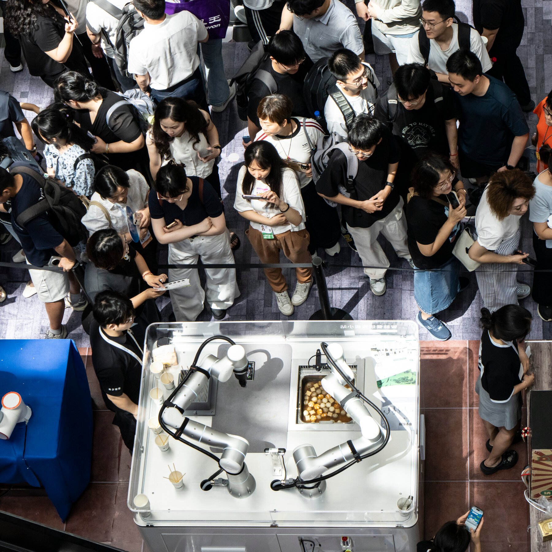 Crowds gather around an automated robotic food preparation station displaying dual robotic arms cooking food in a metal tray.
