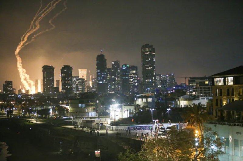 An illuminated coastal city skyline at night with bright streaks of light arcing across the dark sky above lit high-rise buildings and a waterfront promenade.